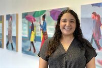 Young woman in dark blouse smiles in front of colorful paintings 